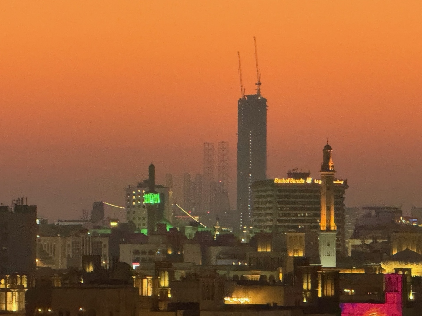 Vibrant dusk cityscape with a skyscraper under construction, illuminated minarets, and traditional architecture under an orange sky.