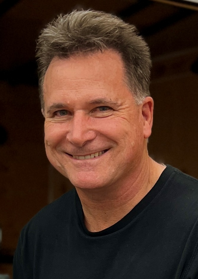 Smiling man with short graying hair and blue eyes wearing a black t-shirt.