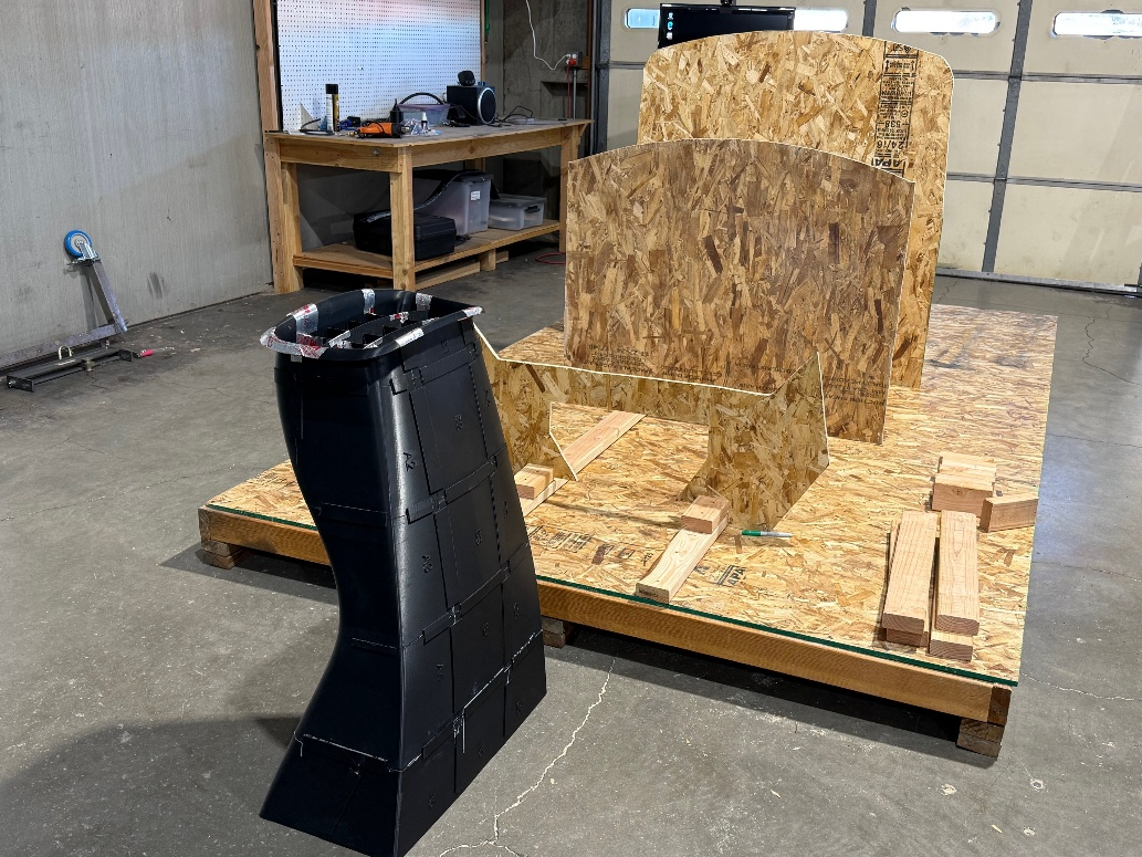 Black curved custom-fabricated component next to an OSB jig with curved panels in a workshop.