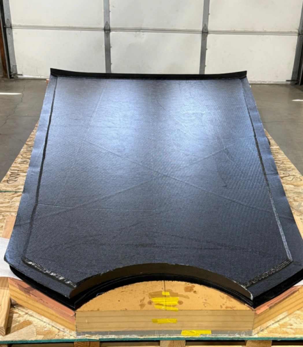 Black woven carbon fiber composite panel with a curved end, resting on a wooden manufacturing mold.