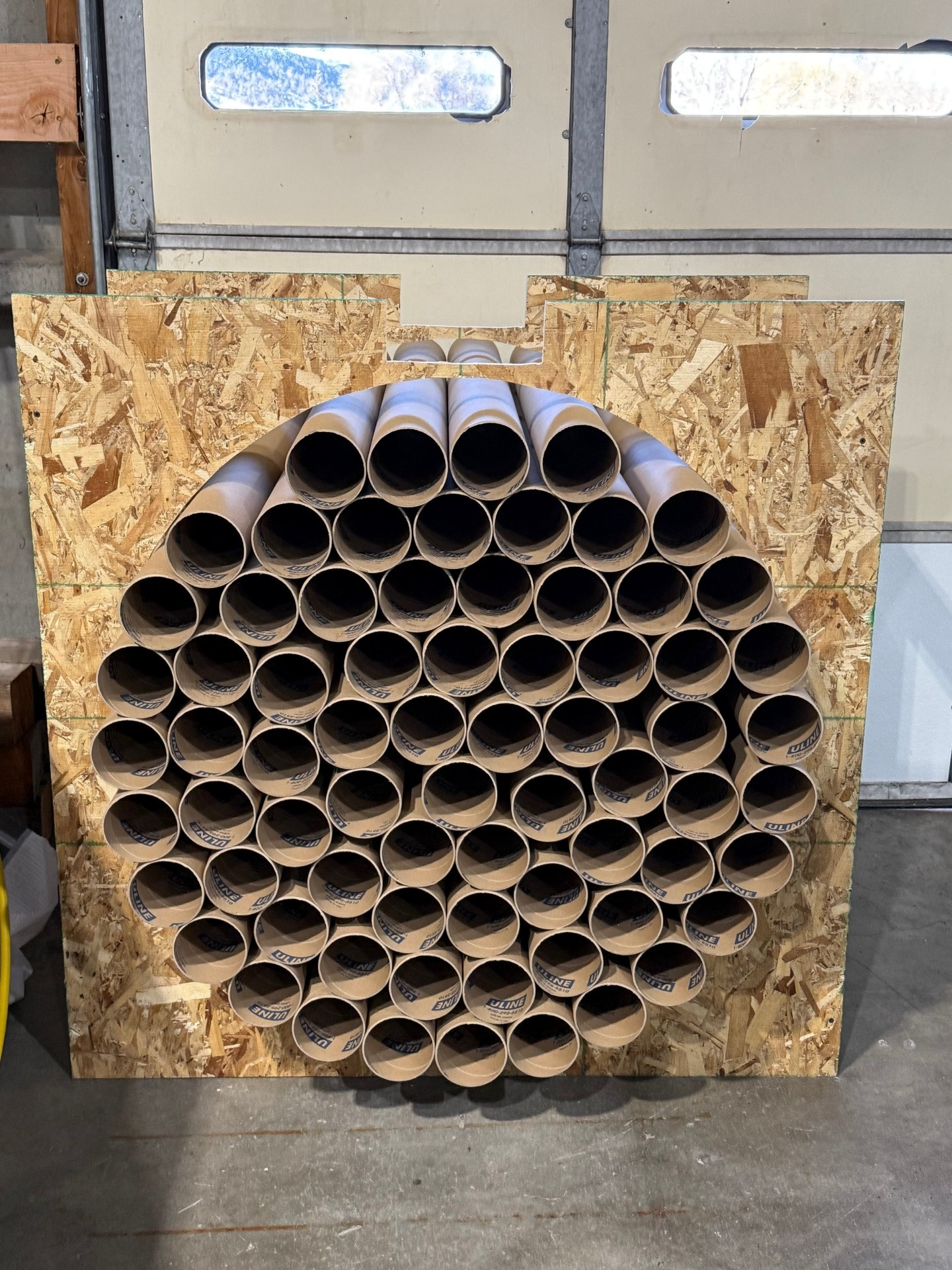 Brown cardboard tubes stacked in a large rounded cutout of an OSB panel, forming a workshop storage rack.