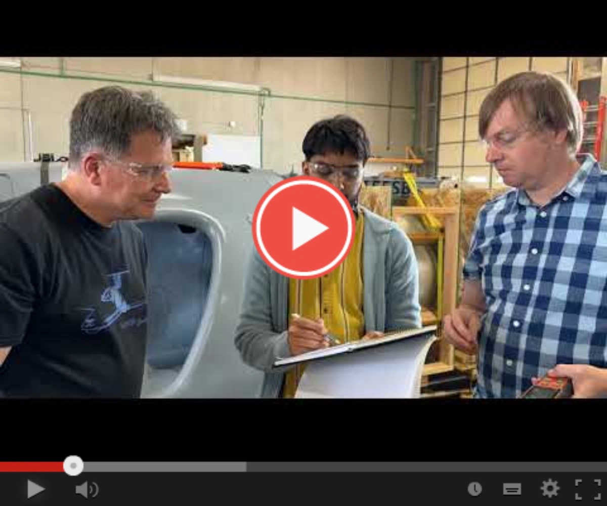 Three men in a workshop review plans for a large white object.