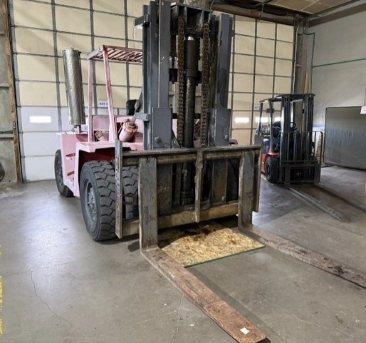 Pink forklift with large forks in warehouse, another forklift in background.